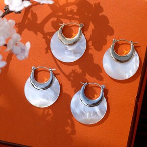 May include: Four pairs of white and gold hoop earrings with a crescent moon shape. The earrings are on a bright orange surface.