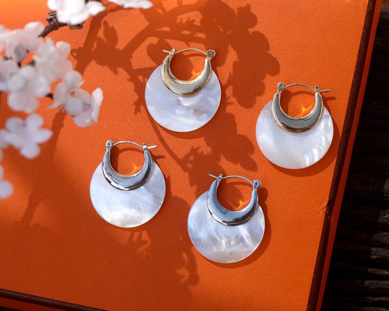 May include: Four pairs of white and gold hoop earrings with a crescent moon shape. The earrings are on a bright orange surface.