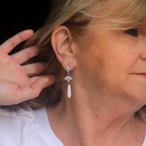 Op de afbeelding: Een vrouw met blond haar draagt een wit shirt en een paar zilveren oorhangers met witte peervormige stenen.