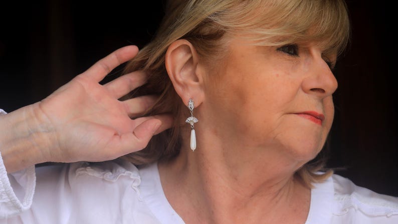 Op de afbeelding: Een vrouw met blond haar draagt een wit shirt en een paar zilveren oorhangers met witte peervormige stenen.