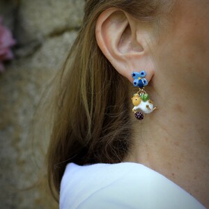 May include: A pair of dangle earrings featuring a white ceramic animal with a green leaf and a blue flower charm. The animal is holding a purple berry.
