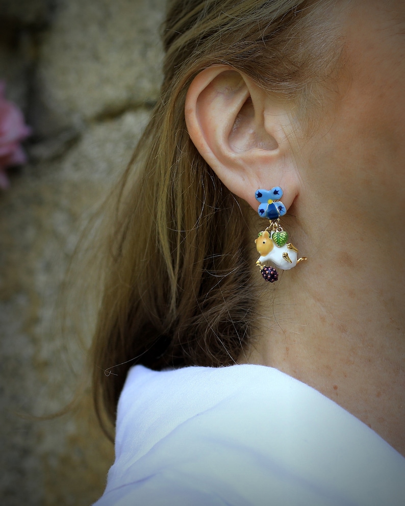 May include: A pair of dangle earrings featuring a white ceramic animal with a green leaf and a blue flower charm. The animal is holding a purple berry.