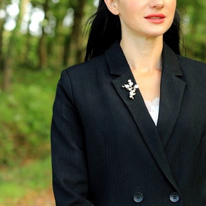 May include: A black blazer with a white pearl and gold brooch. The brooch is shaped like a flower branch.
