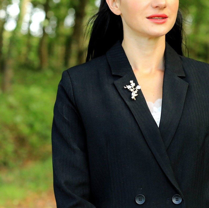 May include: A black blazer with a white pearl and gold brooch. The brooch is shaped like a flower branch.