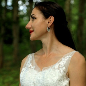 May include: A woman in a white lace dress with a V-neckline stands in a forest. She is looking to the right and has her hair pulled back. She is wearing silver hoop earrings.