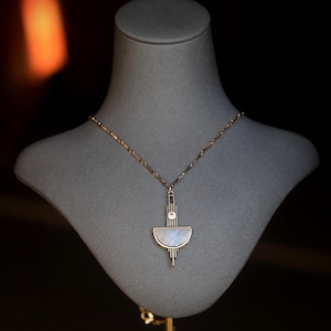 Pode incluir: Um colar de ouro com um pingente geom&eacute;trico em forma de lua crescente com uma pedra transparente no centro. O pingente est&aacute; no pesco&ccedil;o de um manequim.