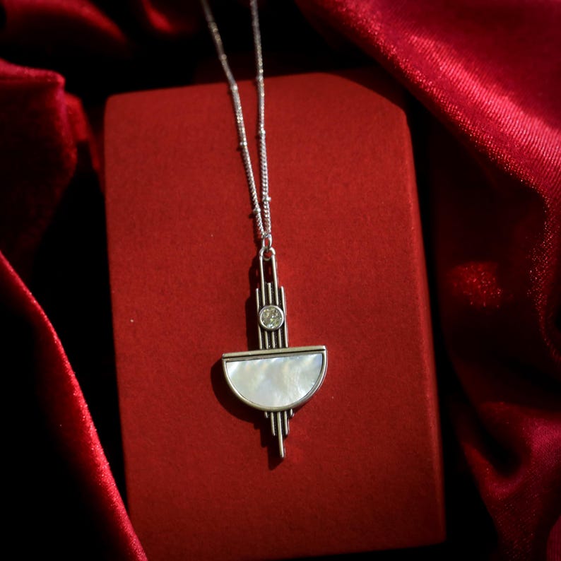 May include: A silver-toned necklace featuring a pendant with a semi-circular white mother-of-pearl inlay, a small circular clear stone, and vertical metallic bars. The pendant hangs from a delicate silver-toned chain, creating a geometric design.
