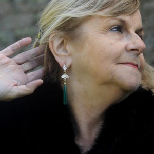 May include: A woman wearing a black fur coat with long, green gemstone earrings. The earrings have a gold setting and a delicate floral design.