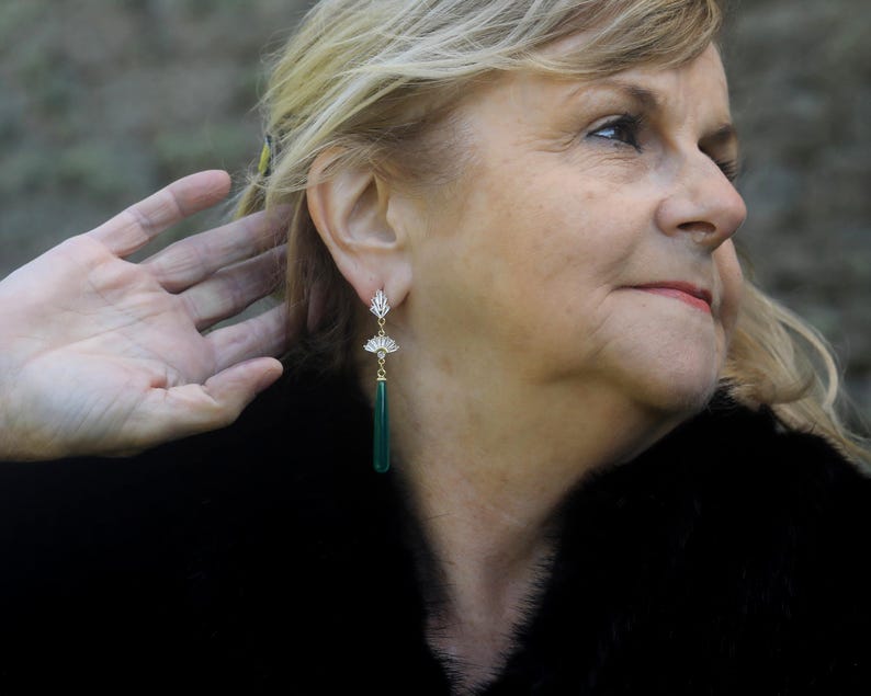 May include: A woman wearing a black fur coat with long, green gemstone earrings. The earrings have a gold setting and a delicate floral design.