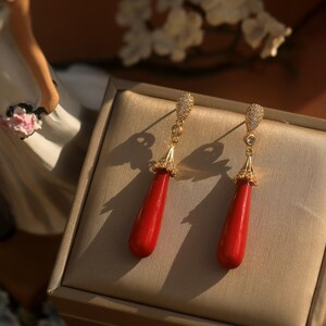 May include: A pair of gold-toned drop earrings with red teardrop-shaped pendants. The earrings have a decorative top with small clear stones and are displayed on a neutral-toned surface.