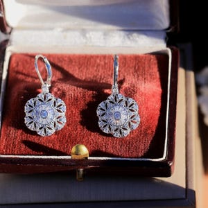 Könnte beinhalten: Ein Paar silberne Ohrhänger mit floralem Design und klaren Steinen. Die Ohrringe befinden sich in einer roten Samt-Schmuckschatulle.