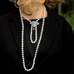 Puede incluir: Un collar largo de perlas en capas con un colgante en forma de abanico. El colgante presenta un diseño geométrico con piedras preciosas verdes y claras. El collar se lleva sobre una blazer negra, creando un look clásico y elegante.