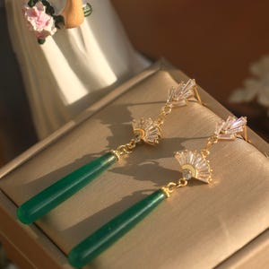 May include: A pair of gold earrings with green gemstone drops and crystal accents. The earrings feature a fan-shaped design with clear crystals.