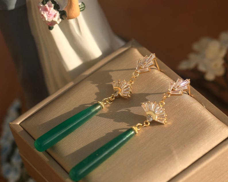 May include: A pair of gold earrings with green gemstone drops and crystal accents. The earrings feature a fan-shaped design with clear crystals.
