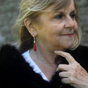 May include: A close-up of a gold earring with a red teardrop-shaped stone. The earring has a small, decorative top with clear stones. The woman in the image is wearing a black fur coat and a white shirt.