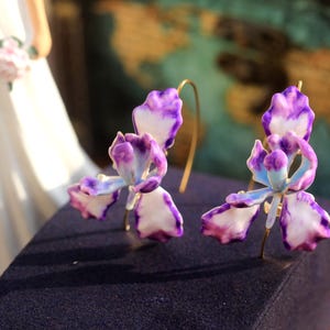 Op de afbeelding: Een paar oorbellen met paarse en witte bloemen met gouden hoepels. De bloemen zijn gedetailleerd en realistisch.