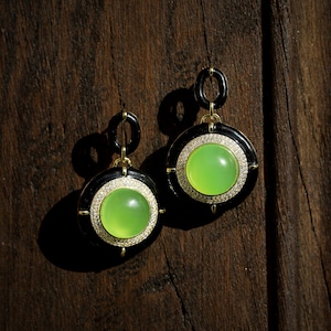 May include: A pair of black and gold earrings with green gemstone cabochons. The earrings feature a round, black enamel setting with a halo of small diamonds surrounding the gemstone.