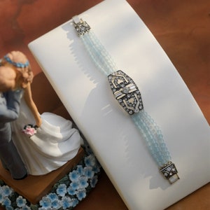 Può includere: Un braccialetto di perline blu chiaro con un pezzo centrale in argento e cristallo. Il braccialetto è esposto su una superficie bianca.