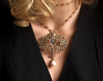 May include: A gold-toned necklace featuring a large, ornate, filigree-style pendant adorned with small clear crystals. The pendant is suspended from a delicate chain and terminates in a single, teardrop-shaped white pearl-like bead at the base.