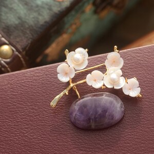 May include: A gold-toned brooch featuring a branch with white flower blossoms and a central oval amethyst stone. The blossoms have a pearl center. The brooch is set against a burgundy leather surface.
