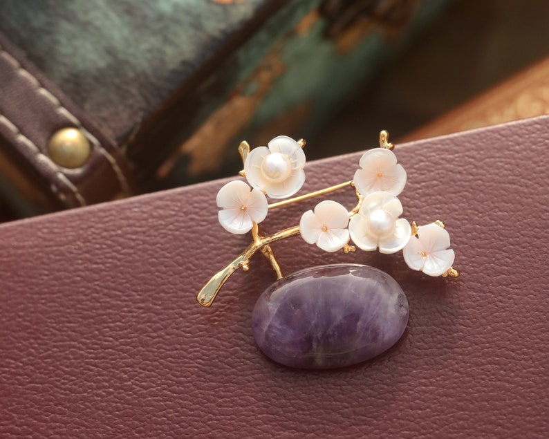 May include: A gold-toned brooch featuring a branch with white flower blossoms and a central oval amethyst stone. The blossoms have a pearl center. The brooch is set against a burgundy leather surface.