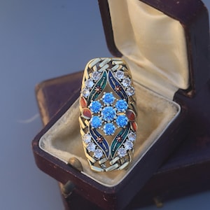 May include: A gold-toned rectangular brooch with a woven design, featuring a central cluster of blue gemstones and surrounding clear gemstones. The brooch is set in a vintage brown jewelry box with a cream-colored interior.