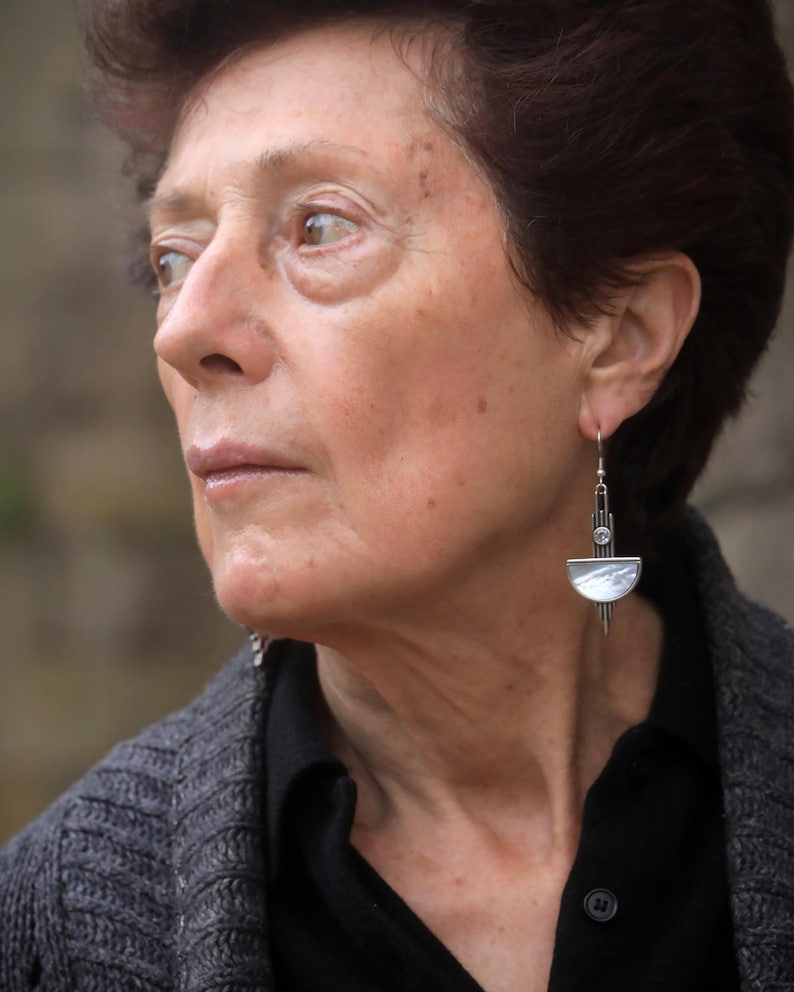May include: A silver-toned drop earring featuring a vertical geometric bar design with a small clear circular stone at the top and a semi-circular mother-of-pearl inlay at the base, suspended from a simple silver-toned hook wire finding.