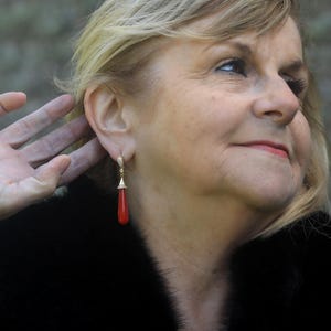 May include: A woman wearing a pair of gold earrings with a red teardrop-shaped coral pendant. The earrings are dangling. The woman is wearing a black fur coat.