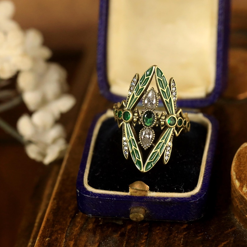 Pode incluir: Um anel de ouro com um design de lib&eacute;lula. As asas da lib&eacute;lula s&atilde;o de esmalte verde com detalhes brancos. O anel tem tr&ecirc;s pedras preciosas em forma de p&ecirc;ra, duas verdes e uma transparente.