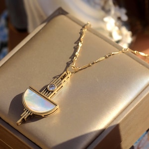 May include: A gold-toned necklace featuring a geometric pendant with a semi-circular iridescent mother-of-pearl inlay, a small clear round gemstone, and intricate linear metal detailing, suspended from a delicate gold-toned link chain.