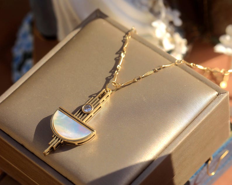 May include: A gold-toned necklace featuring a geometric pendant with a semi-circular iridescent mother-of-pearl inlay, a small clear round gemstone, and intricate linear metal detailing, suspended from a delicate gold-toned link chain.