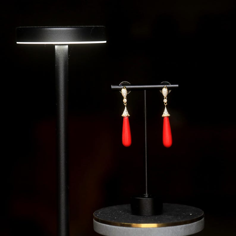 May include: A pair of gold-colored drop earrings with red teardrop-shaped pendants. The earrings are displayed on a black stand against a dark background. The earrings have a decorative top with small, clear stones.