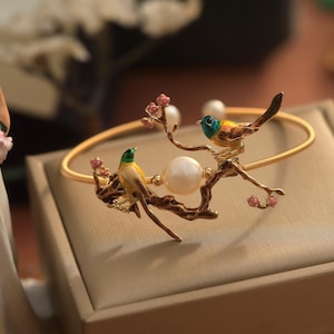 May include: A gold bracelet with a delicate branch design featuring two small birds with green, yellow, and blue enamel. The bracelet has a pearl accent and pink flower accents.