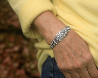 May include: A silver bracelet with a large, rectangular, diamond-encrusted centerpiece. The bracelet is worn on a person's wrist.