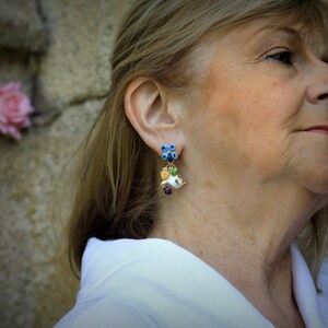 May include: A woman wearing a white shirt with a pair of dangle earrings. The earrings are made of blue, green, white, and yellow glass beads. The earrings are shaped like flowers and have a small, white, bird charm.