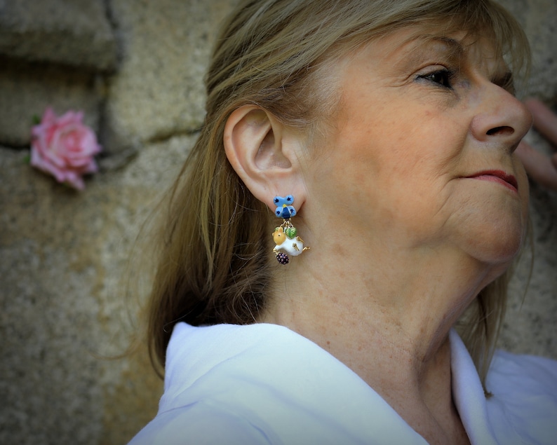 May include: A woman wearing a white shirt with a pair of dangle earrings. The earrings are made of blue, green, white, and yellow glass beads. The earrings are shaped like flowers and have a small, white, bird charm.