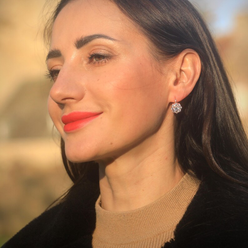 Puede incluir: Una mujer con una chaqueta negra y un su&eacute;ter beige con un par de pendientes de plata colgantes con un grupo de piedras transparentes.