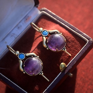 May include: A pair of antique gold-tone earrings with large, round, purple amethyst stones. Each earring features a small, round, blue stone and a long, thin, gold-tone post. The earrings are displayed in a maroon jewelry box.