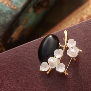 May include: A gold-toned brooch featuring a black oval stone and a floral design. The flowers are white with a pearl center, and the brooch has a pin closure. The brooch is set against a burgundy background.