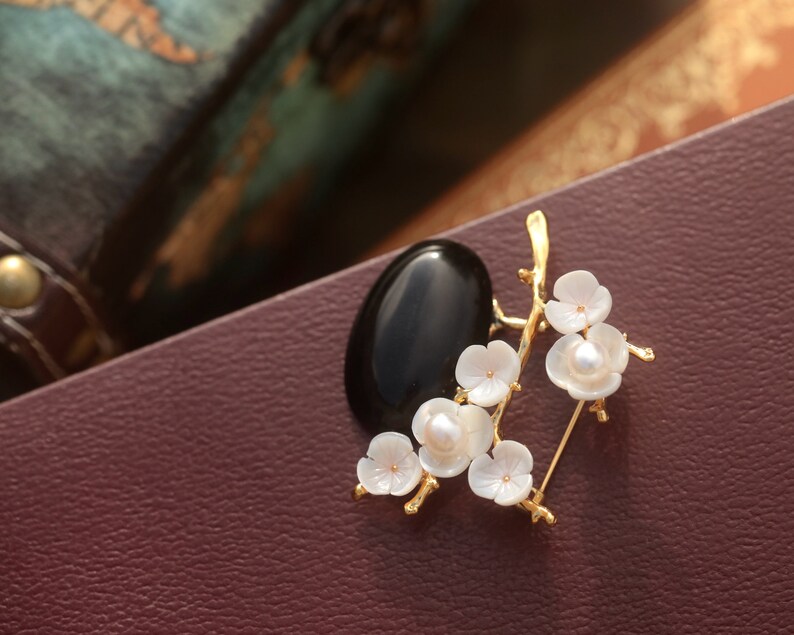 May include: A gold-toned brooch featuring a black oval stone and a floral design. The flowers are white with a pearl center, and the brooch has a pin closure. The brooch is set against a burgundy background.