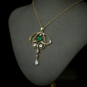 May include: A gold pendant necklace with an Art Nouveau design. The pendant features a central green gemstone, surrounded by clear gemstones and a pear-shaped drop. The necklace is displayed on a gray mannequin.