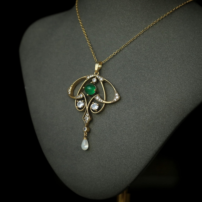 May include: A gold pendant necklace with an Art Nouveau design. The pendant features a central green gemstone, surrounded by clear gemstones and a pear-shaped drop. The necklace is displayed on a gray mannequin.