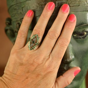 Pode incluir: Um anel de ouro com um design de esmalte verde e pedras preciosas transparentes e verdes. O anel est&aacute; em um dedo com esmalte de unha rosa.