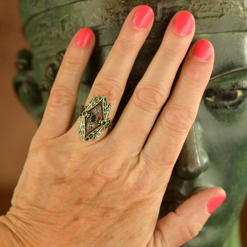 Pode incluir: Um anel de ouro com um design de esmalte verde e pedras preciosas transparentes e verdes. O anel est&aacute; em um dedo com esmalte de unha rosa.