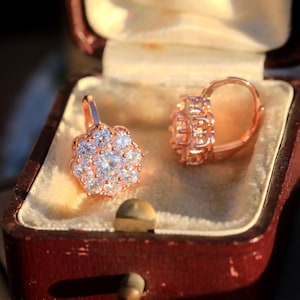 Puede incluir: Un par de pendientes de oro rosa con un racimo de piedras de circonita c&uacute;bica transparentes en forma de flor. Los pendientes est&aacute;n en una peque&ntilde;a caja de joyer&iacute;a de terciopelo rojo.