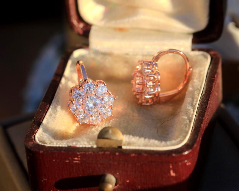 Puede incluir: Un par de pendientes de oro rosa con un racimo de piedras de circonita c&uacute;bica transparentes en forma de flor. Los pendientes est&aacute;n en una peque&ntilde;a caja de joyer&iacute;a de terciopelo rojo.
