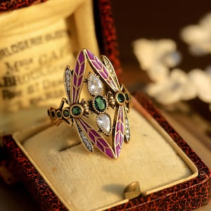 May include: A gold ring featuring a dragonfly design. The dragonfly has purple wings with white and green accents. The ring is displayed in a velvet ring box.