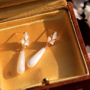 May include: A pair of white pearl drop earrings with gold leaf accents. The earrings are displayed in a brown velvet jewelry box.