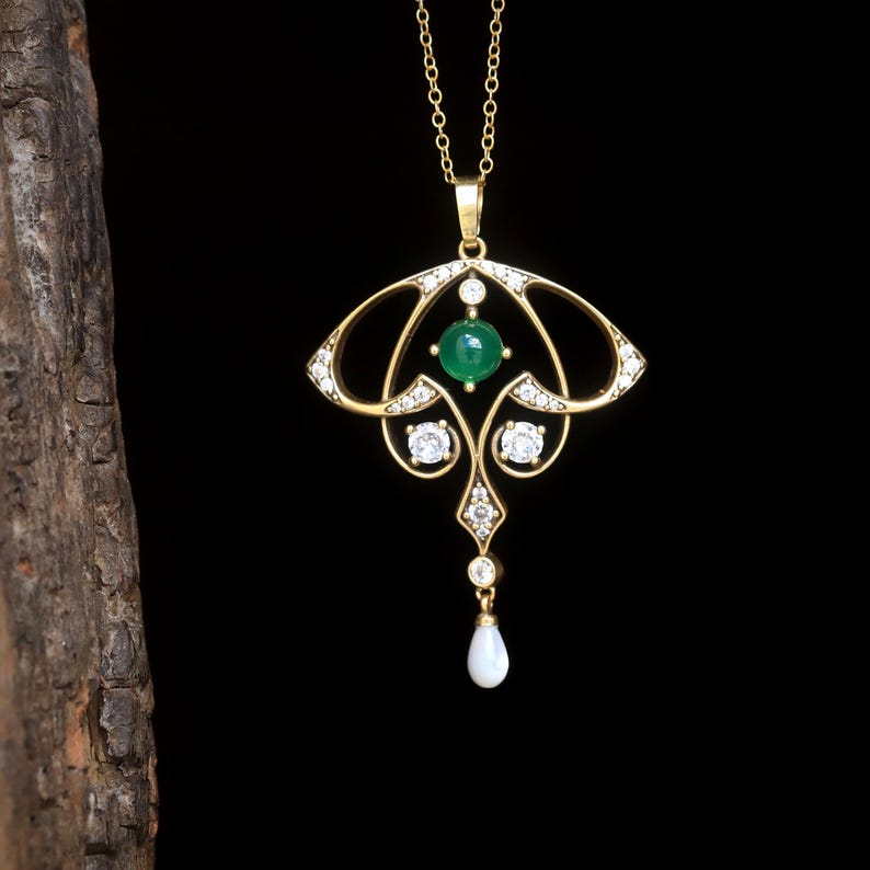 May include: A gold-colored pendant necklace with an Art Nouveau design. The pendant features a central green gemstone, surrounded by clear crystals and a teardrop-shaped pearl. The necklace hangs on a delicate gold chain against a black background.