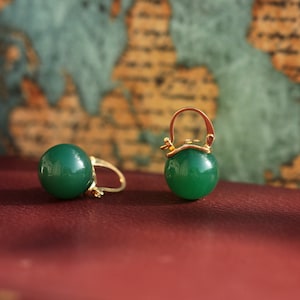 May include: A pair of green, round earrings with gold-colored clasps. The earrings have a glossy finish and a spherical shape. The background features a map design, and the earrings are displayed on a dark red surface.
